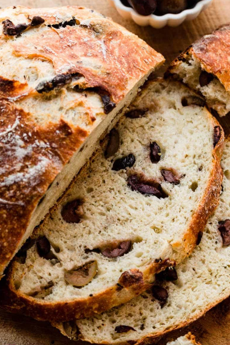 Sliced sourdough bread with Kalamata olives on a wooden board, with a small bowl of olives in the background.